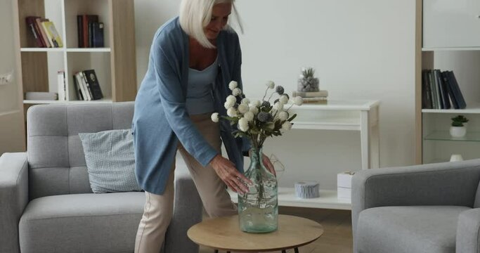 Attractive mature woman carries vase into the living room, puts bouquet of dry pretty flowers on coffee table, decorate domestic room and sits on cozy armchair to rest on weekend leisure alone at home