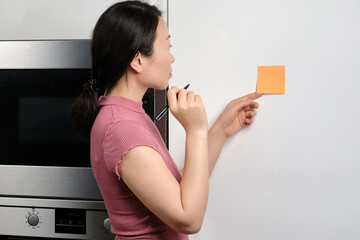 Oriental young woman holding pen and thinking about what to write on generic reminder note stuck onto fridge in house