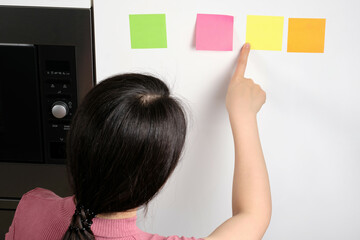 Oriental young woman looking at multicolored generic reminder notes stuck onto fridge in house