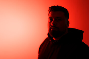 Confident young bearded male model in black hoodie looking at camera while standing in darkness against bright red neon illumination