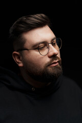 Serious young bearded male hipster in glasses and hoodie against black background
