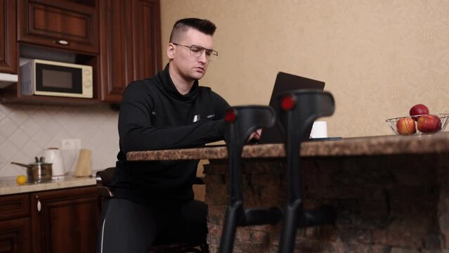 Assistive Technology, Adaptive Equipment, Laptop Work. Bespectacled Man With Leg Prosthesis Works On Laptop, Crutches Beside Him.