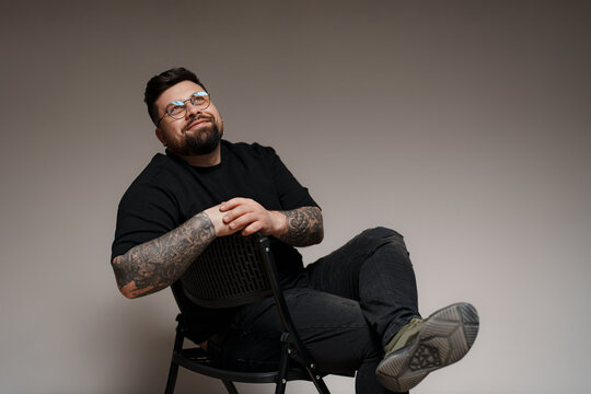 Content young plus size bearded male model in black outfit and eyeglasses with tattooed arms and crossed legs sitting on chair while looking up in studio