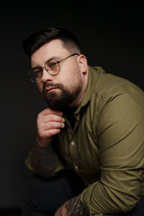 Thoughtful young plump tattooed male model in eyeglasses and green shirt touching beard against black background in studio