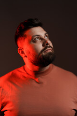 Portrait of pensive young bearded plump male model wearing t shirt against black background with red neon light