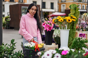 Fototapeta premium A young Latina woman shopping for plants, carrying a basket full of them in a street florist shop.
