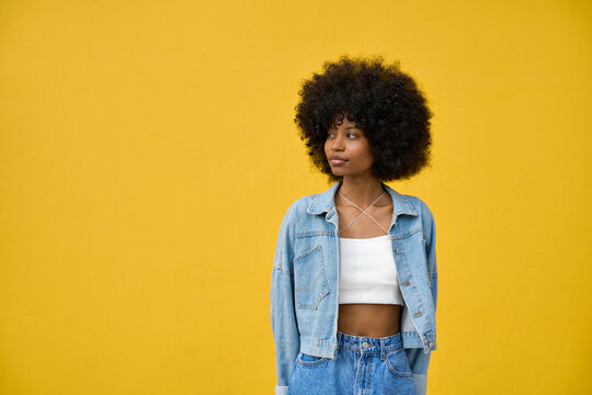 Young stylish woman model pose in a denim jacket in front of a yellow background looking aside. Beautiful afro hair african american girl in trendy clothes stands on orange background with copy space.
