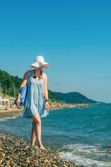 young beautiful woman traveler in a blue summer sundress, sun hat and with a beach bag walks along the seashore during a summer vacation