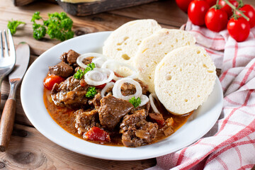 Pork goulash meat with dumplings on white plate, cutlery, garlic, onion, tablecloth in the background - typical Czech food