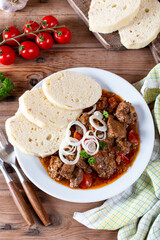 Pork goulash meat with dumplings on white plate, cutlery, garlic, onion, tablecloth in the background - typical Czech food