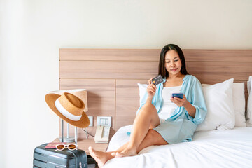 Happy traveler Asian woman holding credit card while using smartphone with travel accessories on bed. Online shopping and travel, cashless society, technology and lifestyle concept. Copy space