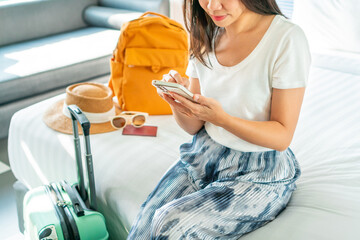 Happy young Asian traveler woman using mobile phone while relax on bed in hotel room. Female plan a trip on cellphone at home. Travel alone, Planning for summer holiday concept