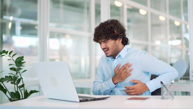 Sick young man suffering from chest pain while working on laptop while sitting at workplace at desk in modern office. Worker keeps his hand on his chest. He has a heart attack. Medical help is needed