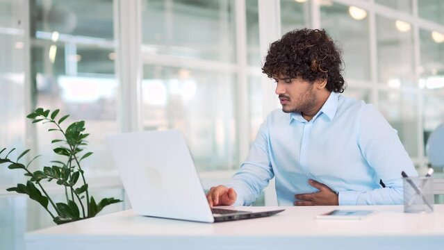 Sick young man suffering from stomach pain while working on laptop while sitting at workplace at desk in modern office. The worker has digestive problems, cramps, food poisoning or constipation