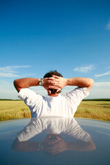 Man Leaning on Car Overlooking Sky and Farmland with clasped hands behind head.