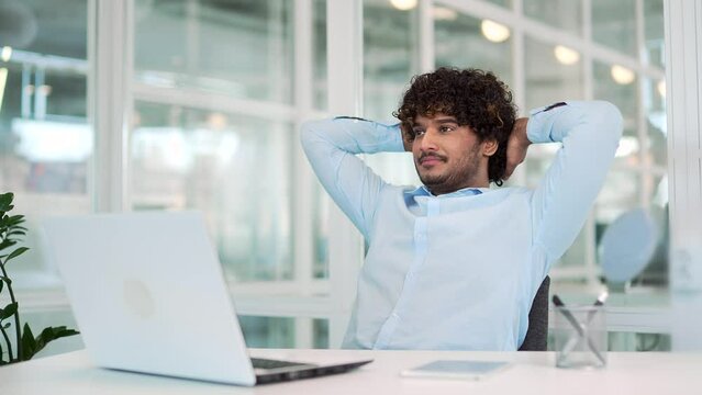 Young happy worker stretching on chair, relaxing with hands behind head with eyes closed, sitting at workplace in modern office. Smiling handsome developer taking a break from working on a laptop