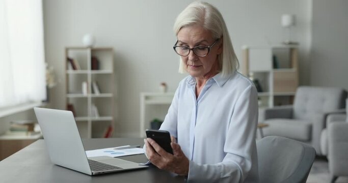 Mature businesswoman sit at desk, hold smartphone, type message, share SMS, browse website, look through information, use new mobile business, professional, financial apps. Workflow use modern tech