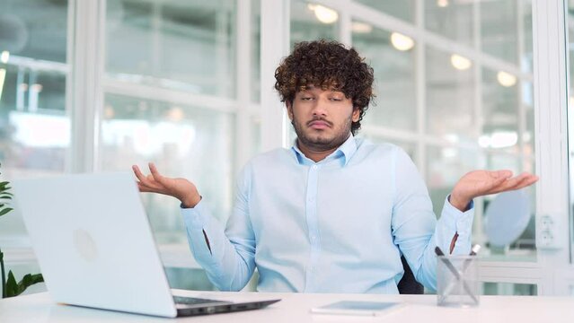 Young surprised puzzled worker spreads hands, shrugs shoulders looking at camera working on laptop while sitting at workplace at desk in office. Man in the shirt has a confused expression on his face