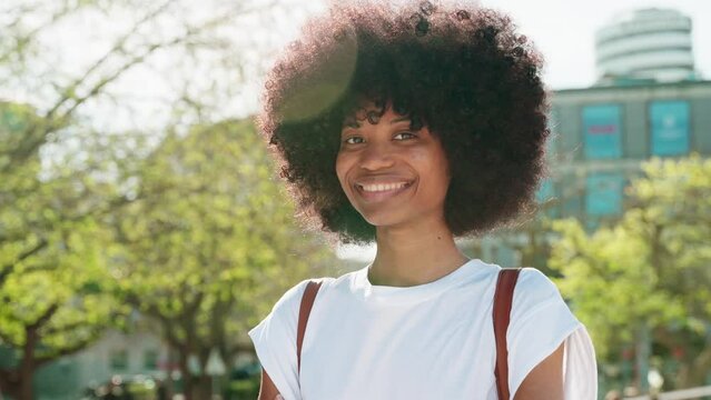 A portrait of African American girl with curly afro hairstyle stands with backpack and holds exercise books in university campus. Smart student looking aside on blurry building background, copy space.