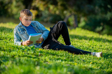 Schoolboy with a textbook in his hands is lying on a green lawn in a park during summer day. Studying teen looks to the notebook in a garden. Pupil reading his notes and memorizing the text outdoors