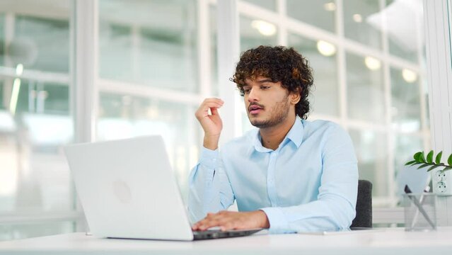 Tired young employee is bored working on laptop while sitting at workplace at desk in modern office. Overworked employee cannot concentrate on work. A lazy male in a shirt is distracted from the task