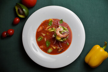 Shrimp with cherry tomatoes, avocado, cucumbers, and radishes in a red sauce on a white plate against a dark green background.