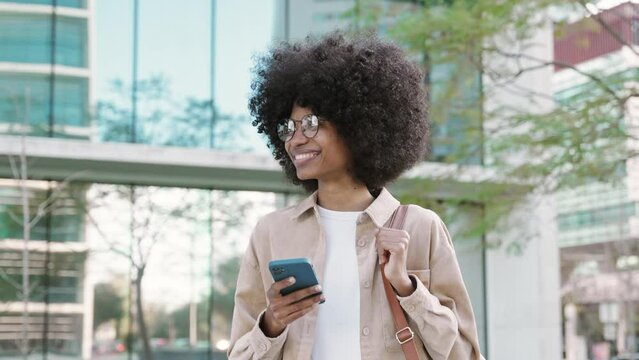 Happy African American student girl with backpack walking outdoors in city and enjoy online communication, scroll social media app on cell phone device on university campus background.