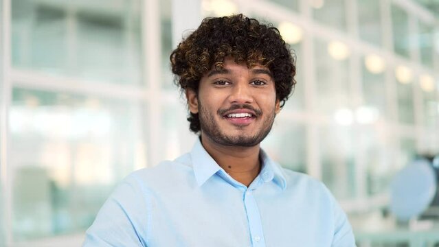 Close up portrait of young smiling handsome man in shirt standing in modern office. Happy male Indian worker or businessman with curly hair with a friendly look looking at the camera. Head shot