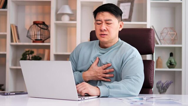 Chinese freelance businessman feeling unwell experiencing panic attack having difficulty breathing, accompanied by chest pain. Man touches chest in attempt to soothe himself while alone in office. 