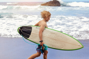 Fit young surfer man with curly blond hair with surfboard goes by the ocean having fun doing...