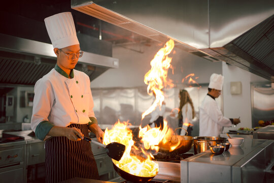 Cook cooking in kitchen with burning pan. Chef in the restaurant kitchen making flambe on frying pan. Cooking food in a frying pan with fire. Chefs doing flambe on food..