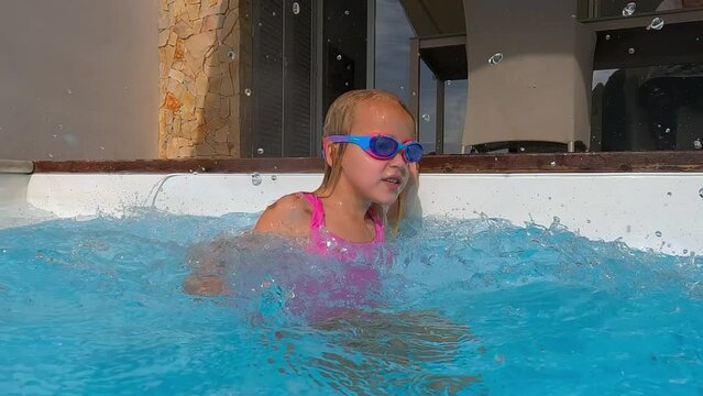 Slow-motion video of a girl in a pink swimsuit and blue swimming goggles playfully splashing water in a pool, enjoying the flying droplets, sun, summer, and vacation