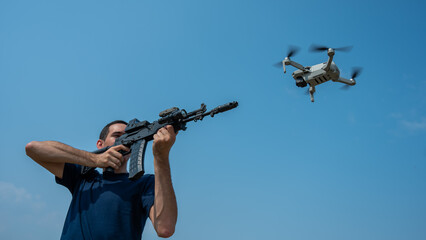 A man aims to shoot a rifle at a flying drone against a blue sky. 