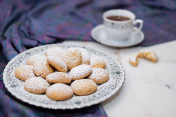Kahk with sugar sprinkled on top, in a rounded china plate and a cup of tea, on white marble background, (Kahk is a traditional cookies after Ramadan for eid)

