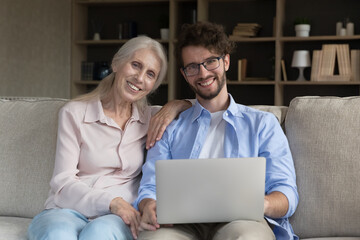 Cheerful smart young man helping with computer to senior mom. Happy adult son and elderly mother using laptop on home sofa together, holding gadget, looking at camera, smiling