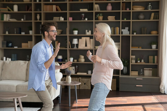 Cheerful energetic old retired mom and active adult son guy enjoying movement, fitness activity at home, dancing to music together in modern living room, smiling, laughing