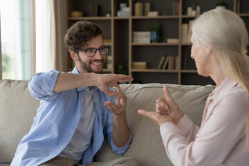 Cheerful adult son talking with hands to senior old mom with hearing disorder, disability, using...