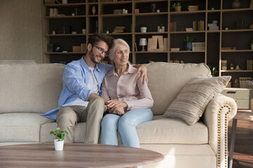 Young adult grandkid visiting elder grandma at home, hugging granny. Positive senior mother and son...