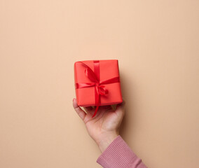 A woman's hand holding a red gift box wrapped with ribbon on a beige background, a concept of congratulations, surprise