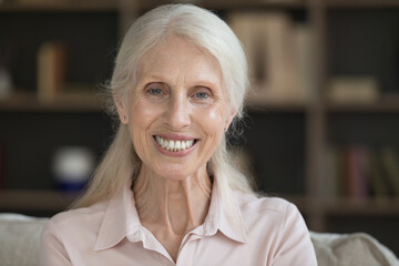 Happy positive retired elderly lady home head shot portrait. Cheerful senior woman looking at camera with toothy smile, posing indoors. Video call screen view