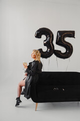 a blonde in a black with champagne on the couch. balloons of numbers thirty-five