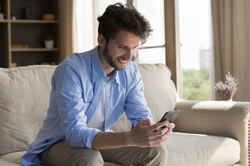 Joyful happy handsome young man using online application, media service on smartphone, enjoying connection, Internet communication at home, holding mobile phone, looking at screen, smiling