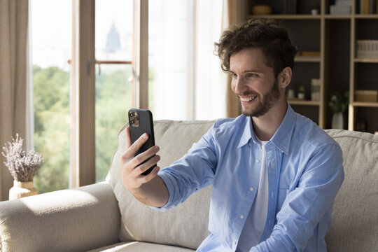 Happy handsome young blogger guy taking selfie on mobile phone, talking on online conference call, shooting head shot video for blog, holding smartphone in outstretched hand