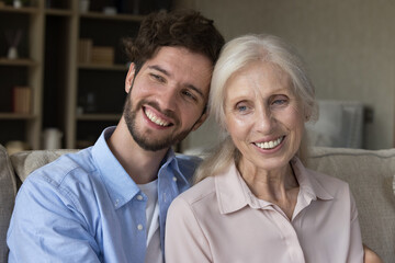 Happy grandson guy and positive older grandma sitting close on home sofa, hugging, looking away with toothy smiles. Family portrait of handsome adult son and senior mom