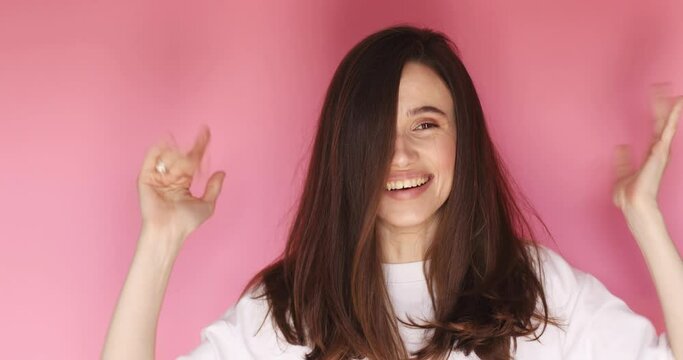 Cheerful happy joyful young woman, brunette loose hair cheerfully throws. Cute girl hands raised. Smiling face. Happy woman fix hair, look funny and funky isolated on pink background.