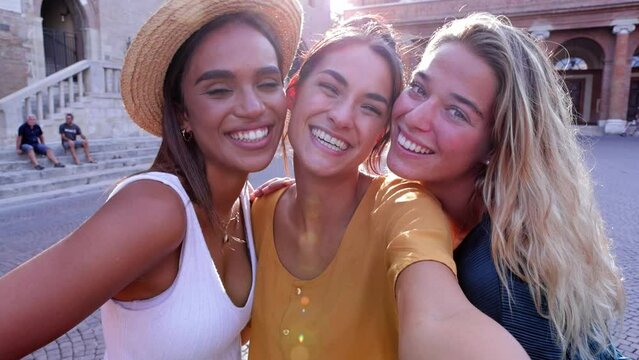 Three united diverse young female friends smiling at camera together. Multiracial pretty women hugging each other laughing while taking selfie video portrait during summer vacation. Friendship concept