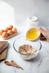 Glass mixing bowl with various baking ingredients around it. Ingredient is poured into bowl and mixed by hand.
