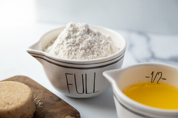A ceramic measuring bowl filled with wheat flour sits on a white marble surface next to melted butter and a wooden cutting board with packed brown sugar