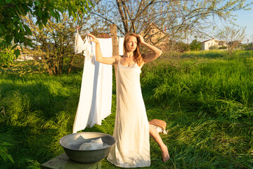beautiful red girl in nightie hanging laundry outdoors. village woman working in countryside.Cute girl in dress washing white clothes in metal basin in backyard, hanging laundry on clothesline 