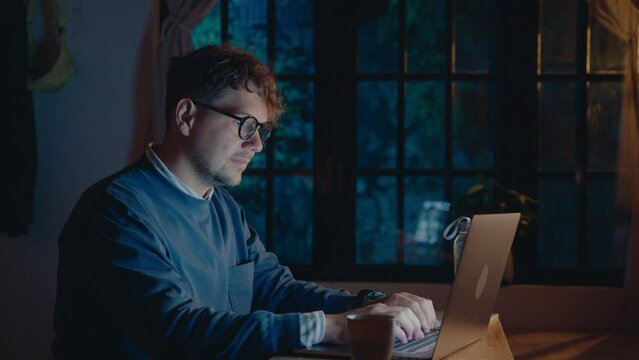 Young man in eyeglasses typing on laptop in dark room, working remotely overtime during night. Medium shot